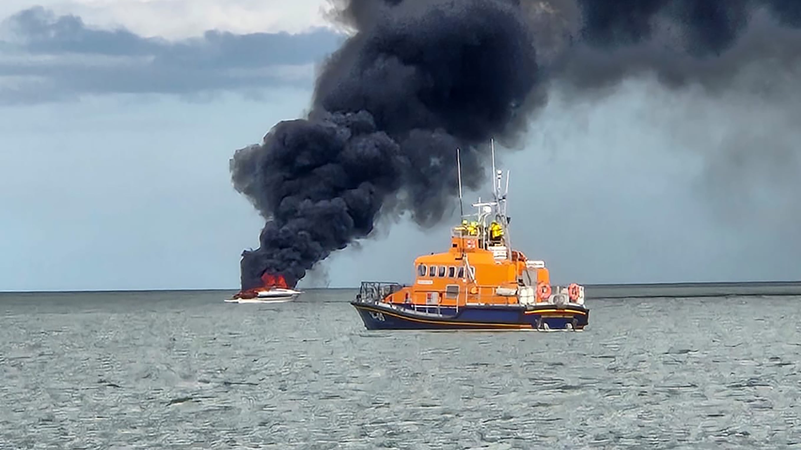 An all-weather lifeboat at sea travelling from right to left, with a white motorboat in the background giving off plumes of black smoke. Photo: Brian Darcy 