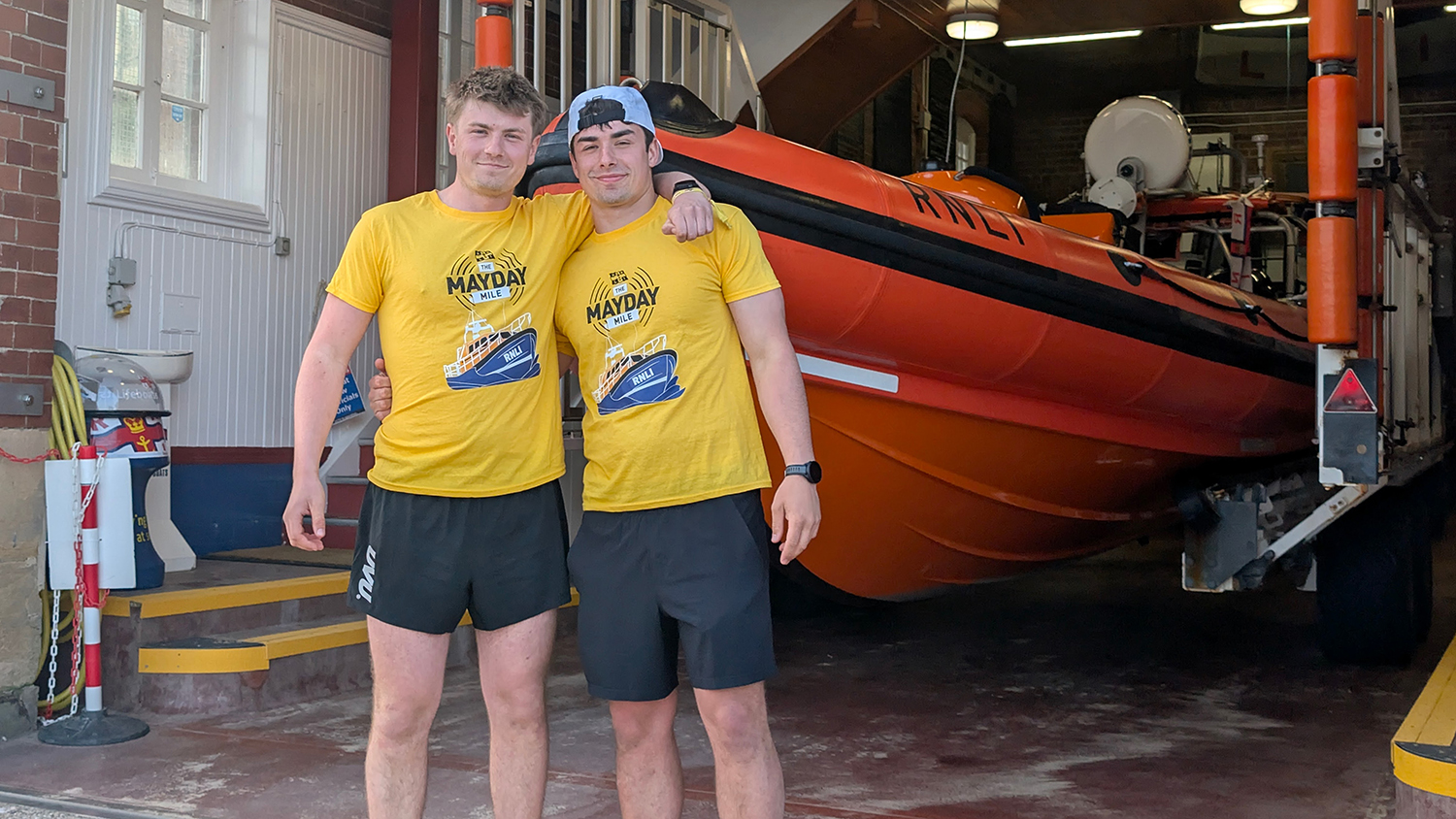 Brothers Euan and Andrew Gray are standing in front of Cullercoats Lifeboat Station. They have their arms around each other’s shoulders and are smiling at the camera. They are wearing bright yellow RNLI Mayday Mile T-shirts, black shorts and yellow trainers.
