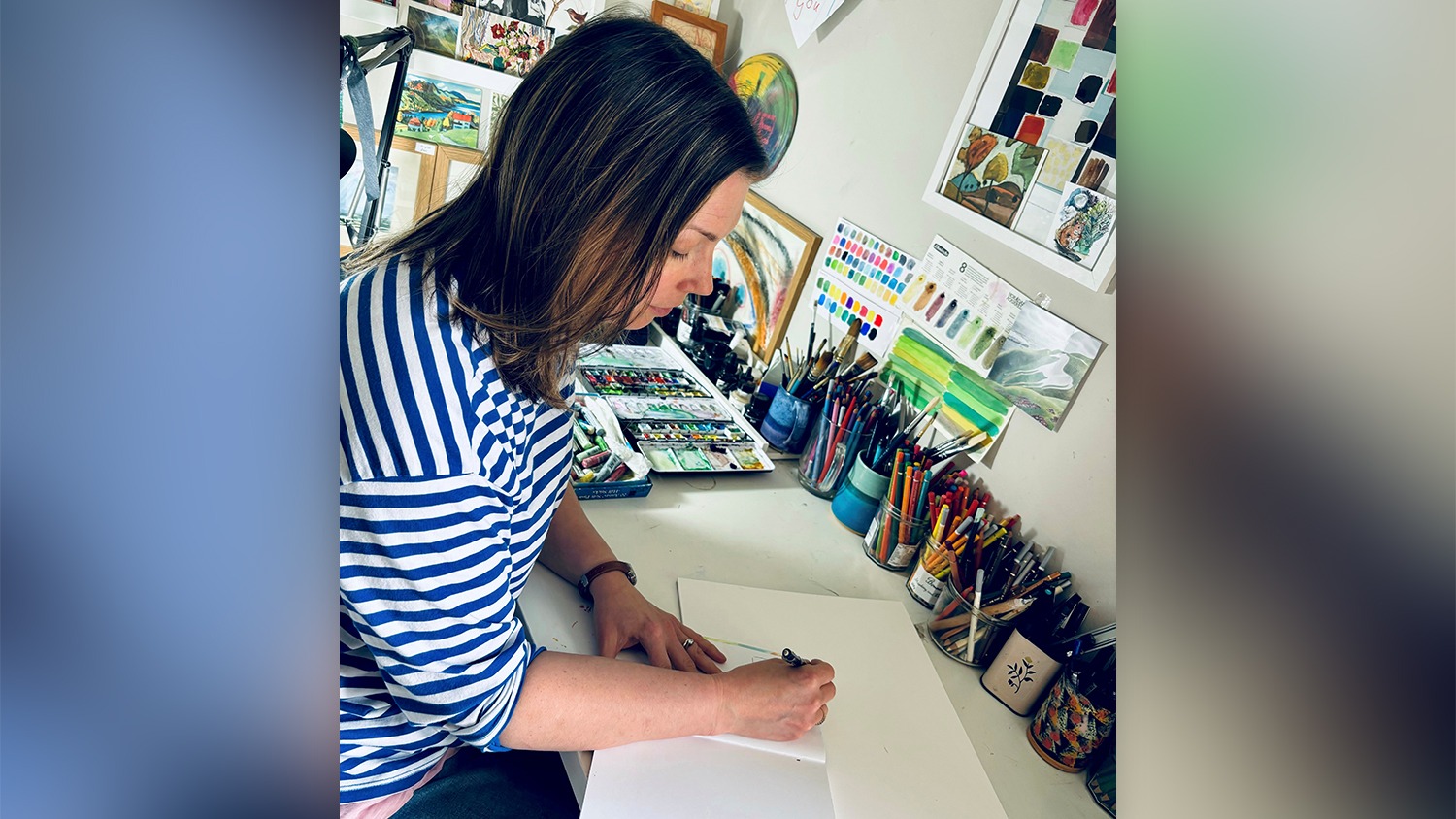 Hannah Lawson is sitting at a desk in her art studio. She is surrounded by art supplies, including pots of paintbrushes, pencils, pens and paints. There is lots of art on the walls. She is sketching in a notebook and is wearing a blue-and-white stripe top.
