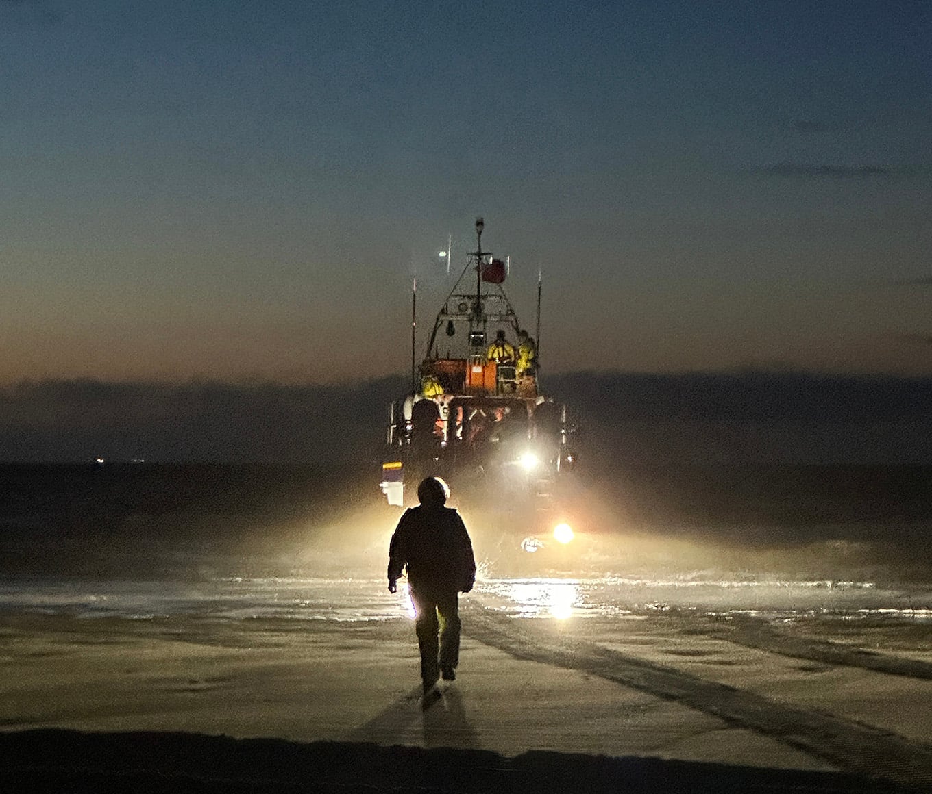 Hoylake RNLI's Shannon class all-weather lifeboat launches from its launch and recovery system at dusk. A strong beam of light from the recovery system is shining onto the shore.