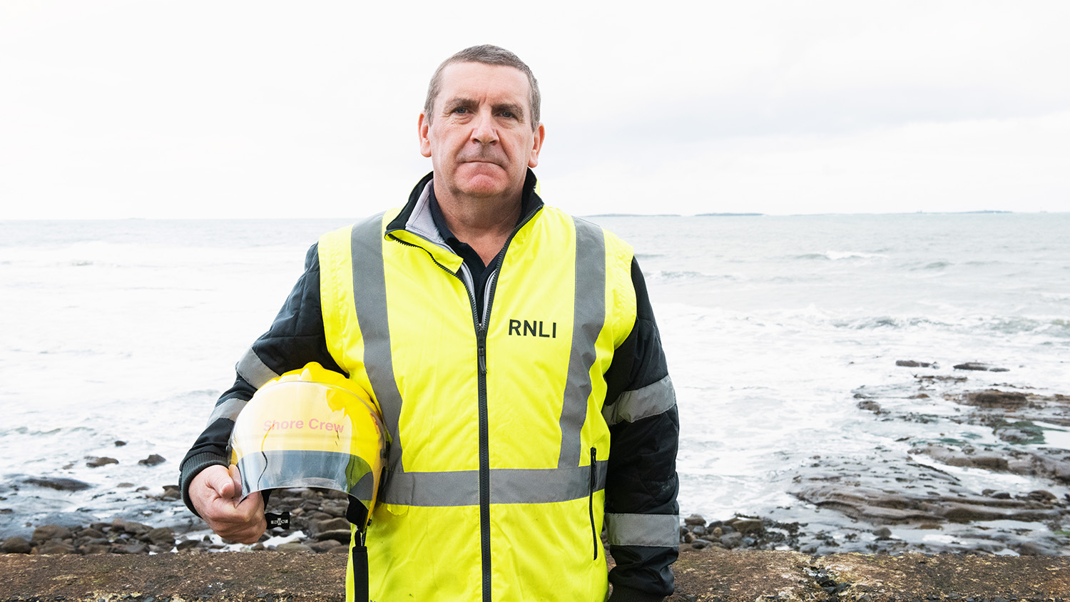 Lee stands on the shoreline, wearing a high-vis jacket and holding a helmet.