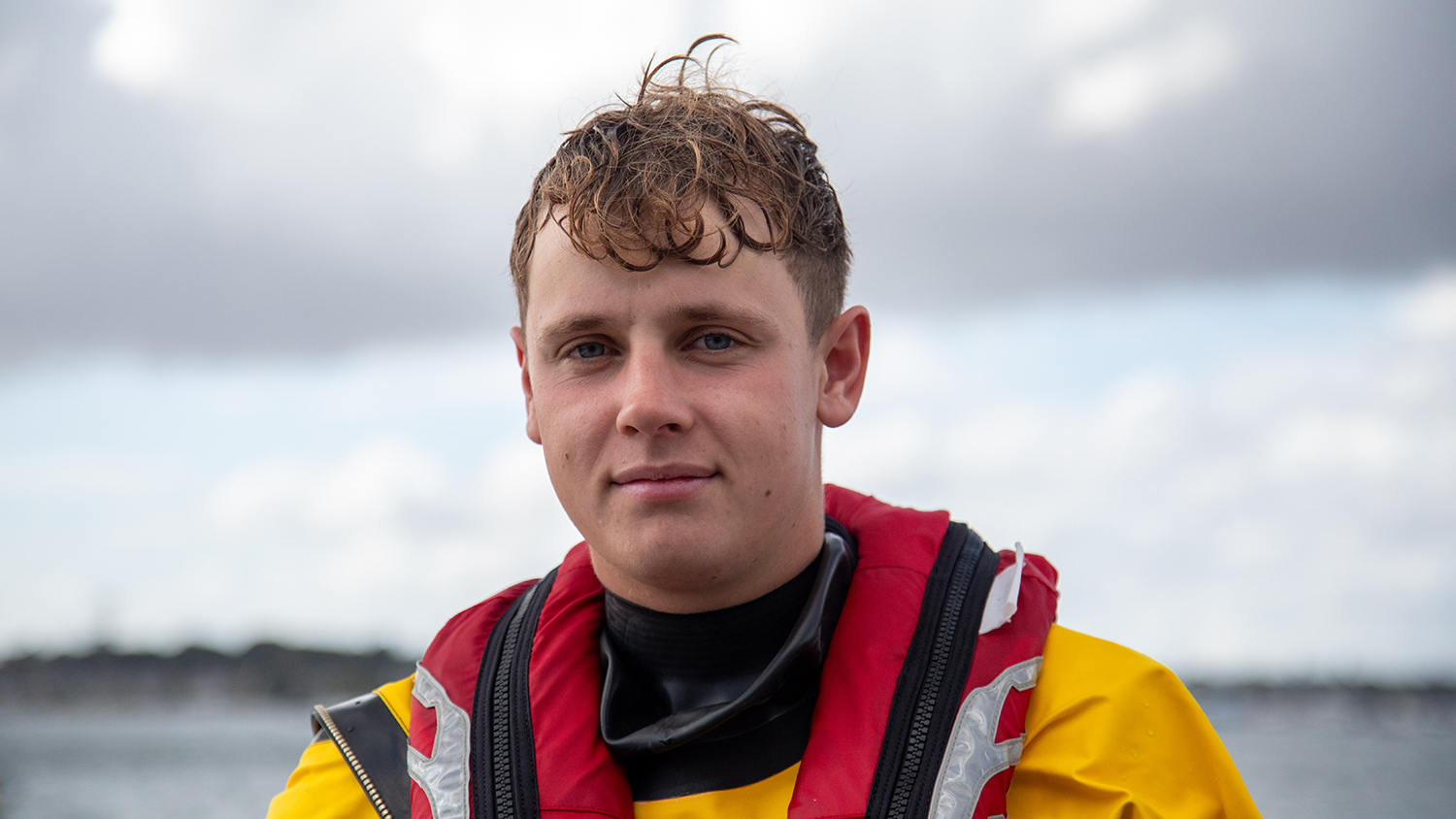A head and shoulders shot of Burnham-on-Sea RNLI Volunteer Lifeboat Crew Member Jake Coombes in crew kit with the sea in the background.