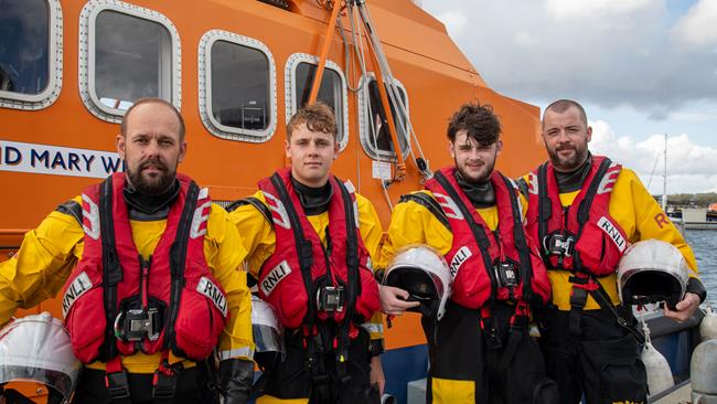 Four volunteer lifeboat crew members – two fathers and their sons – standing in crew kit in front of an all-weather lifeboat at the RNLI College in Poole, Dorset. Photo: RNLI/Charlotte Hill.