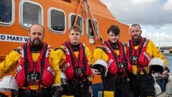 Four volunteer lifeboat crew members – two fathers and their sons – standing in crew kit in front of an all-weather lifeboat at the RNLI College in Poole, Dorset. Photo: RNLI/Charlotte Hill.