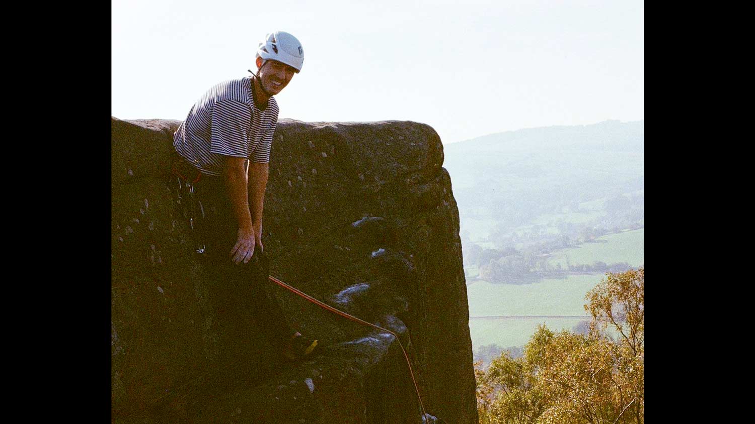 A man rests against the top of a large rock wearing climbing gear. He is wearing a white helmet and has chalk on his hands to help his grip. Behind him, green hills roll into the distance.