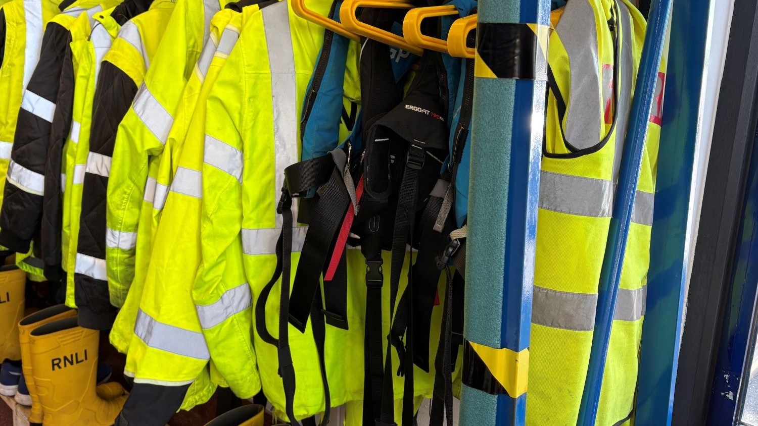 Fluorescent yellow jackets, blue lifejackets and hi-vis vests hanging on a rail with yellow wellies on a shelf underneath.
