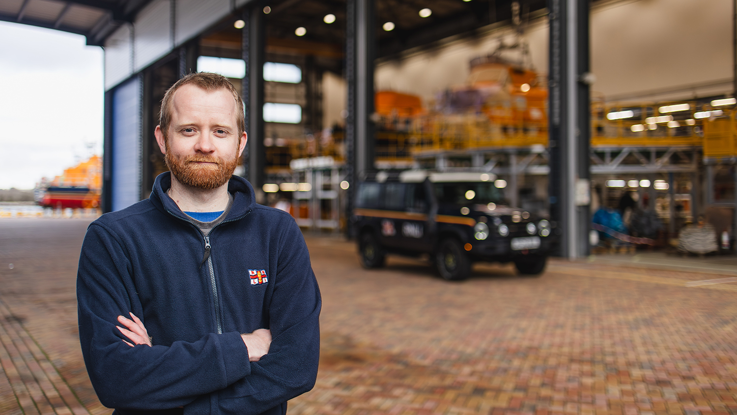 RNLI Engineer Sam Barton facing the camera with his arms folded. He has a neutral expression with a slight smile. He is wearing an RNLI-branded zip-up fleece. Sam is standing outside the All-weather Lifeboat Centre in Poole, Dorset, and an INEOS Grenadier is parked up behind him. 