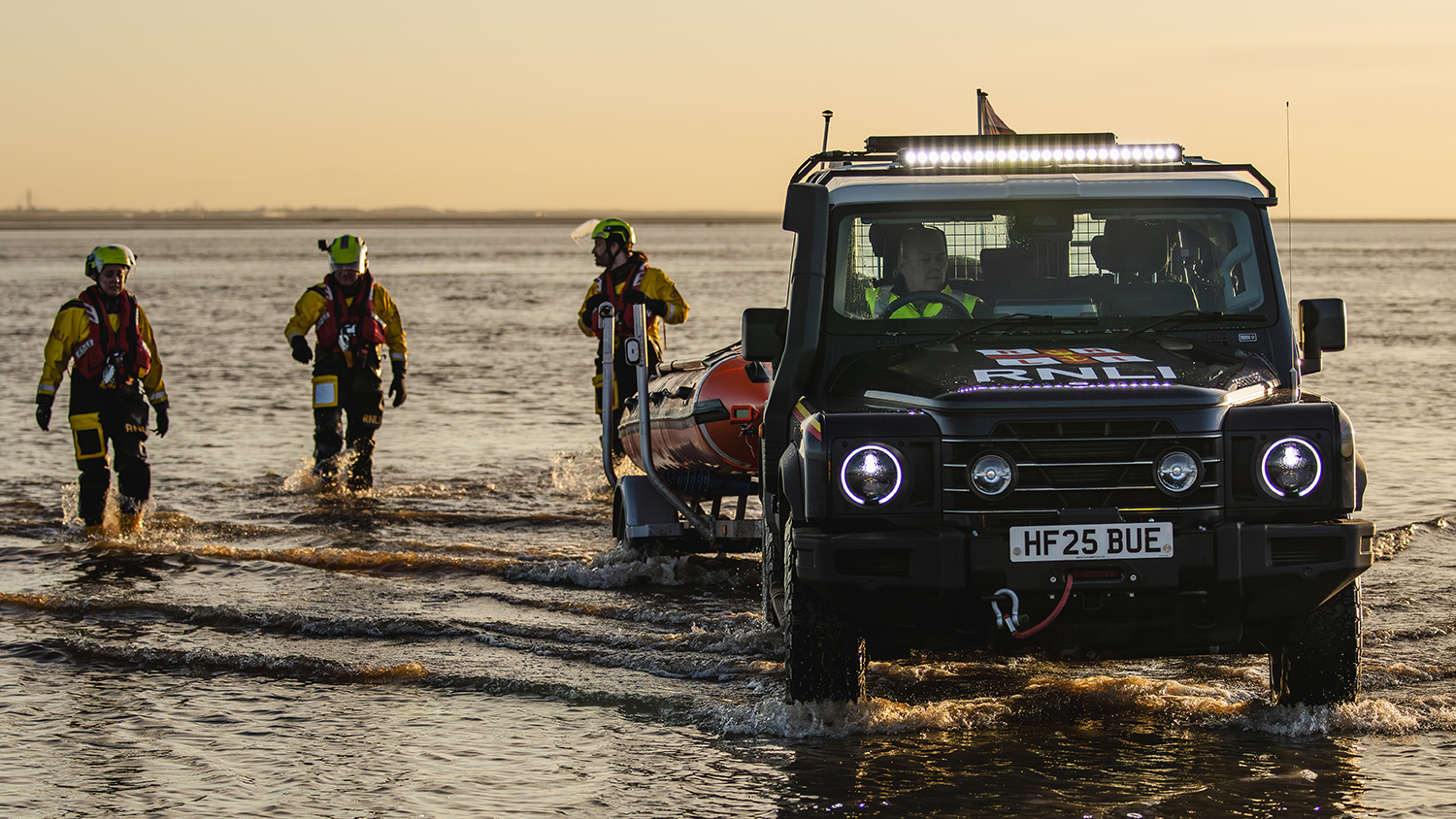 The Grenadier at Lytham St Annes is in the water, with their inshore lifeboat in a trailer attached to the vehicle. Three crew members are wading through the shallow water. The roof lights and headlights on the Grenadier are switched on. The sun is setting and the sky is a golden yellow colour. 