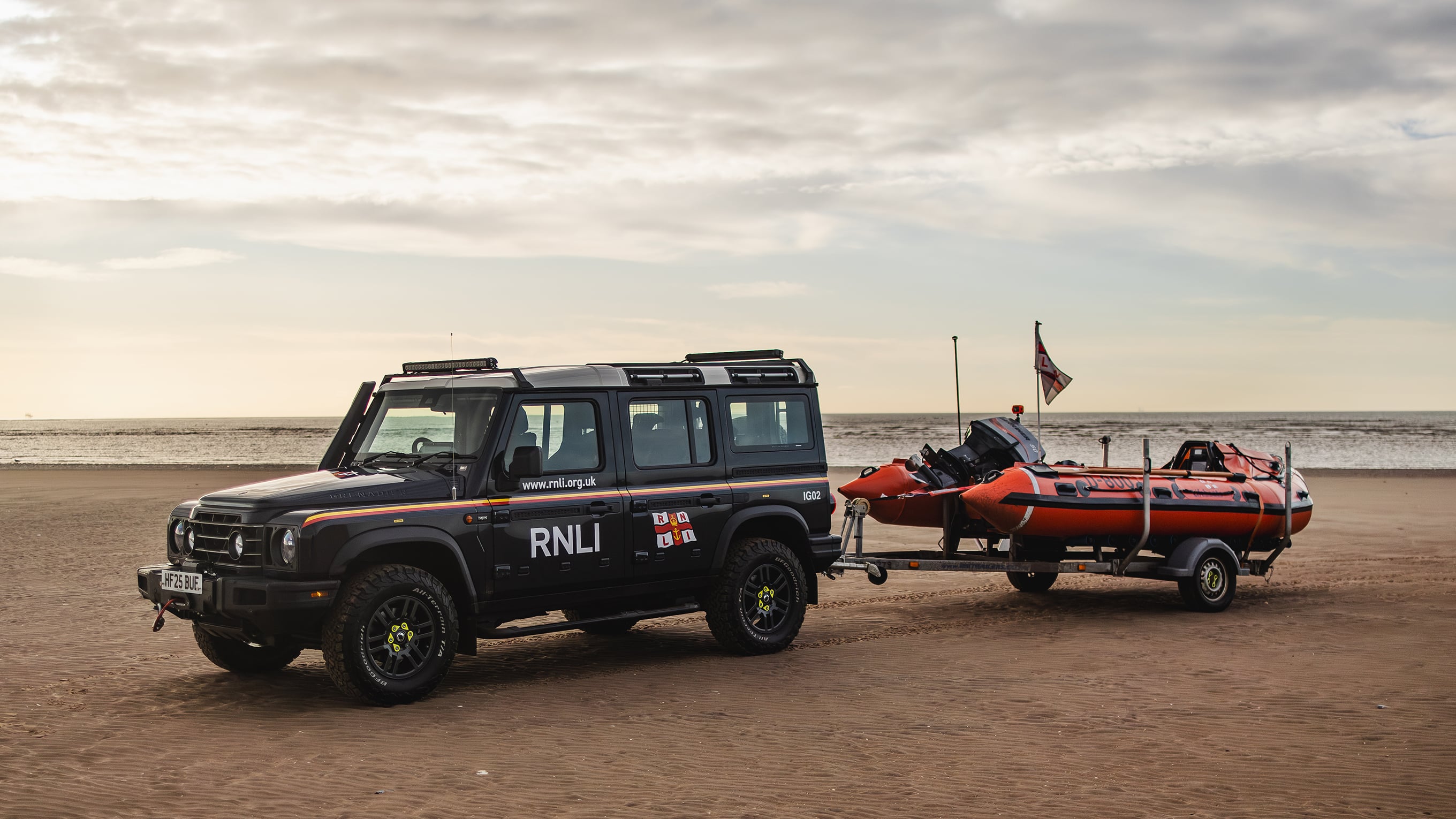 An INEOS Grenadier vehicle is parked on a sandy beach. The Lytham St Annes D class lifeboat is on the back of a trailer, which is attached to the Grenadier. 