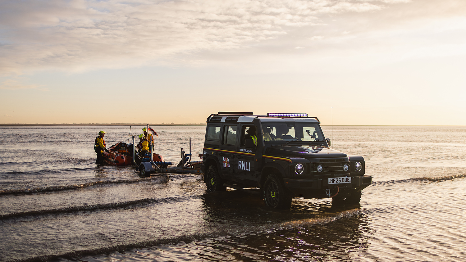 An INEOS Grenadier vehicle is in calm, shallow water at the beach in Lytham St Annes. The Lytham St Annes D class lifeboat is on the back of a trailer, which is attached to the Grenadier. There are three crew members in the water with the lifeboat. The sun is setting in the sky. 