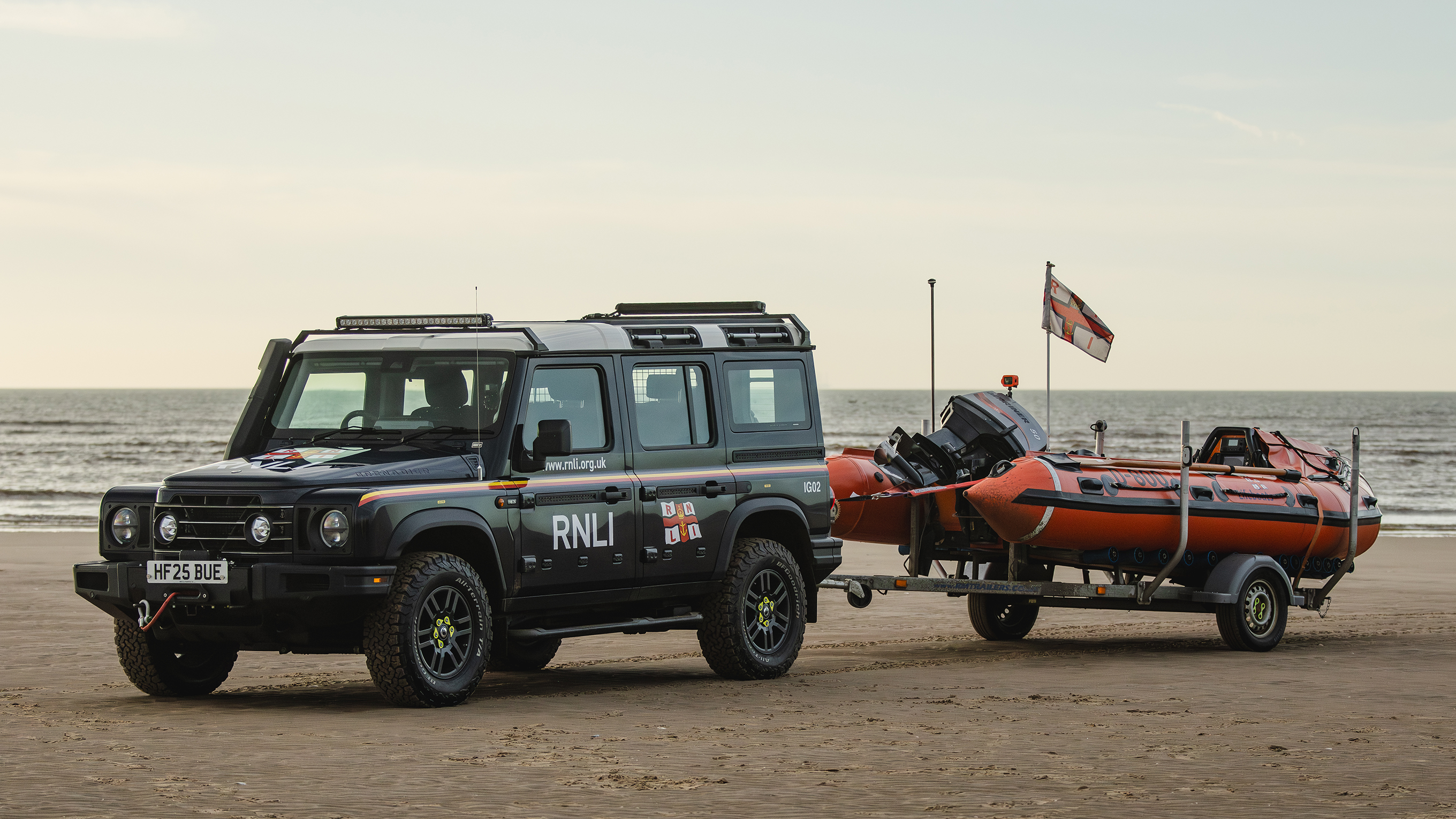 An INEOS Grenadier vehicle is parked on a sandy beach. The Lytham St Annes D class lifeboat is on the back of a trailer, which is attached to the Grenadier. 
