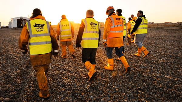 A group of shore crew in yellow wellies and hi-viz walk across shingle at sun set towards the boathouse.