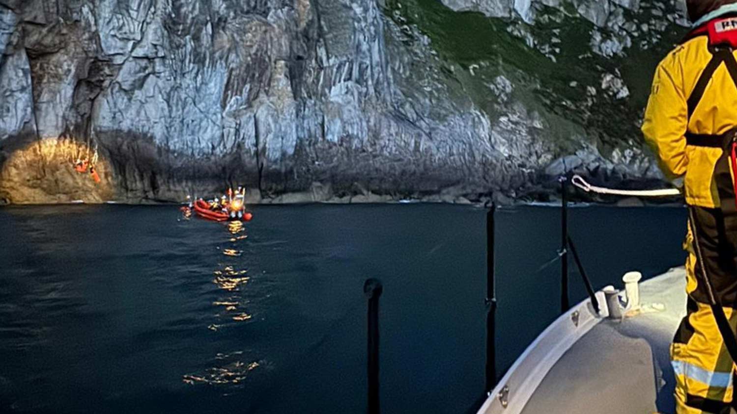 A lifeboat crew member onboard the deck of an all-weather lifeboat watches on as a B class lifeboat floats on the water, near a cliff. A spotlight illuminates Coastguard volunteers at the cliff’s base, tending to the casualty.