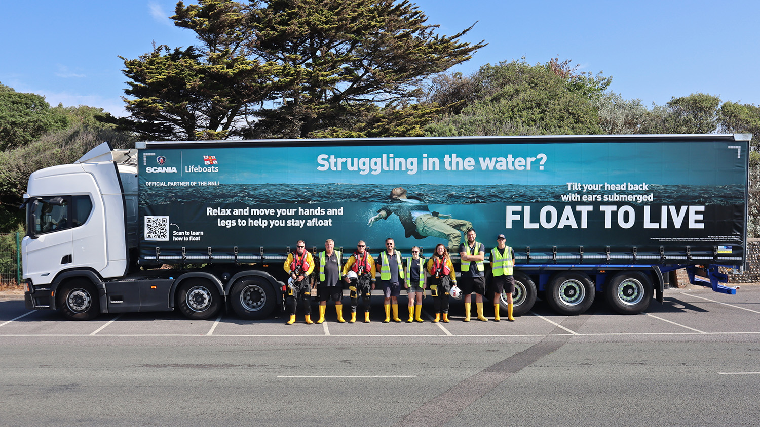 A Scania trailer, with the RNLI’s Float to Live advert across the side. Littlehampton RNLI volunteers are standing in front of the truck. 