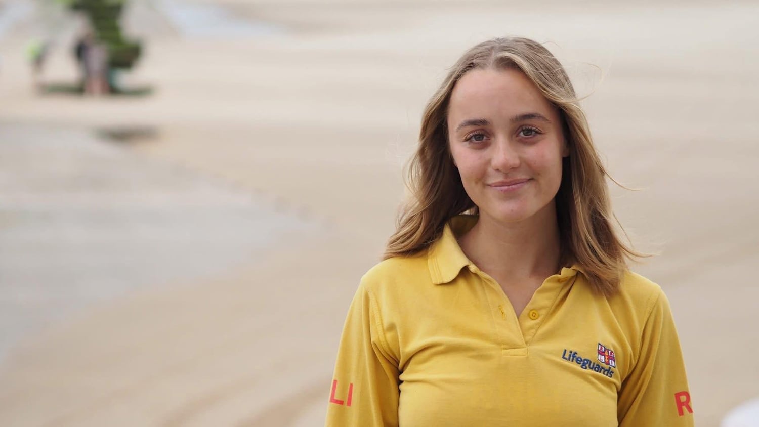 Lois Kemp, an RNLI Senior Lifeguard, smiles at the camera. She’s wearing a yellow RNLI lifeguards polo top and standing in front of a sandy beach.