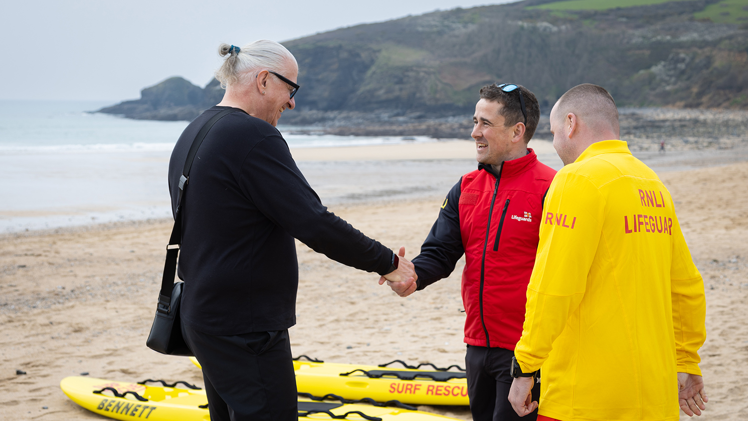 Rescuee Mark on the beach, shaking hands with an RNLI lifeguard as another lifeguard looks on.