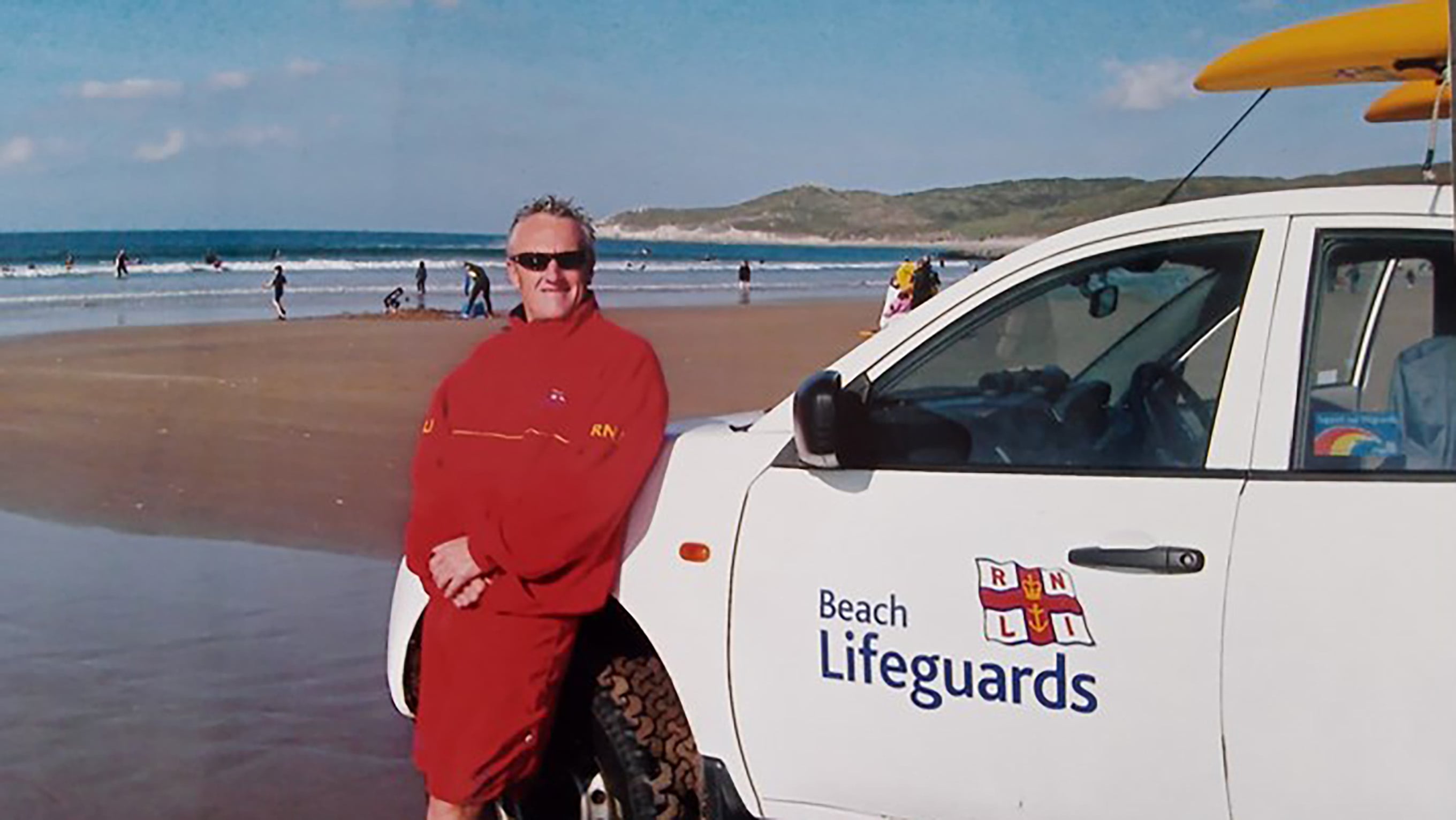 Nik, as an RNLI lifeguard. He’s wearing a red fleece and shorts and leaning against an RNLI patrol vehicle.