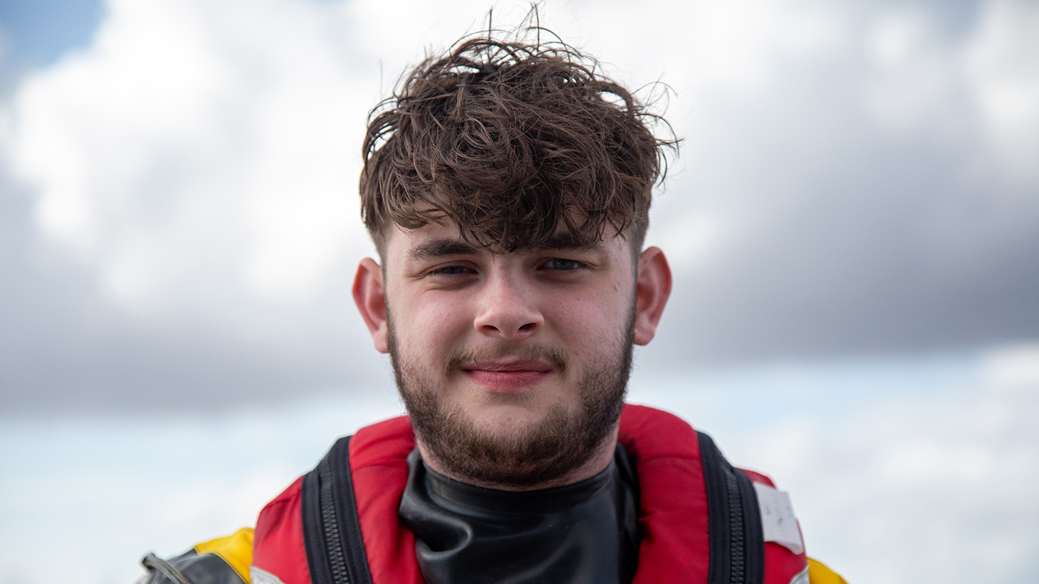 A head and shoulders shot of Rhyl RNLI Volunteer Lifeboat Crew Member Rhys Jones in crew kit with the sea in the background.