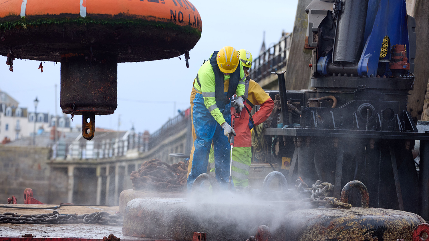 A large metal sinker is being jetwashed by a man in wearing high-vis gear and a yellow hardhat. 