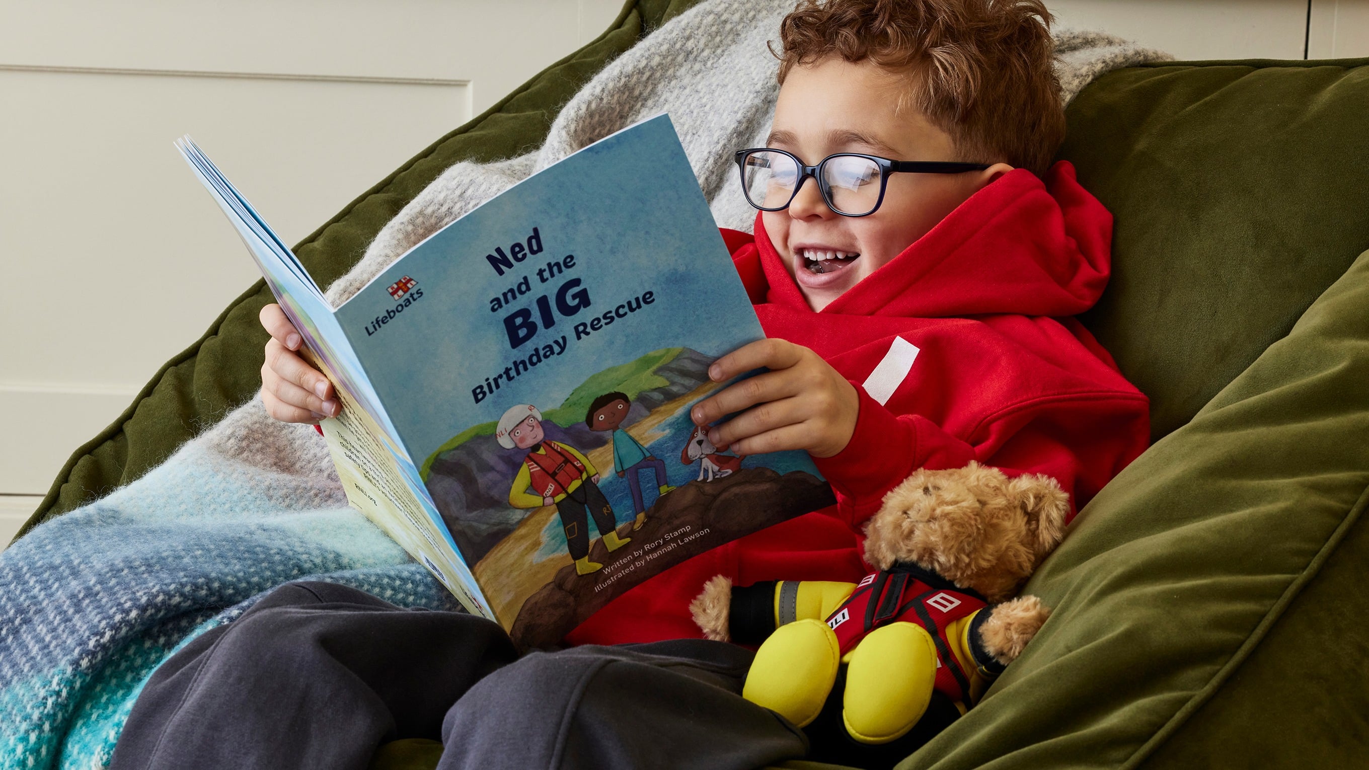 A child sitting on a beanbag reading The Big Birthday Rescue personalised book. The child is wearing glasses and a red hoodie, and is holding the book with both hands. He is smiling at the book. On the beanbag is a blue blanket and an RNLI lifeboat crew teddy bear. Photo: CHS Agency.