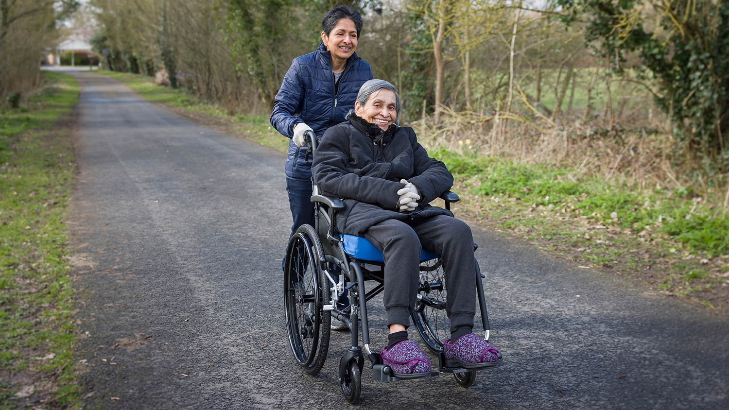 A woman pushing an older woman in a wheelchair down a coutnry road. They're both smiling. 