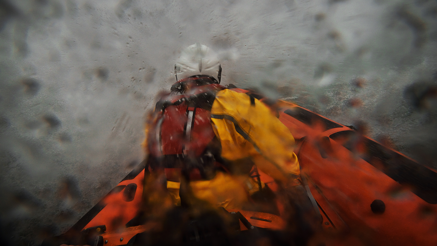 A crew member in an inshore lifeboat, surrounded by sea spray and rain.