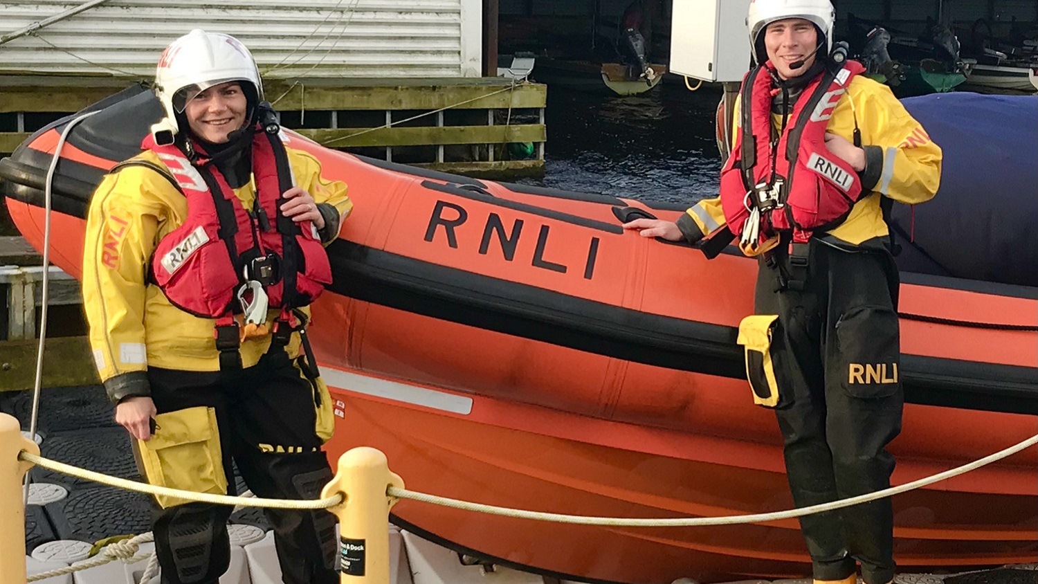 One male and one female RNLI crew member stands and smiles in full RNLI kit.