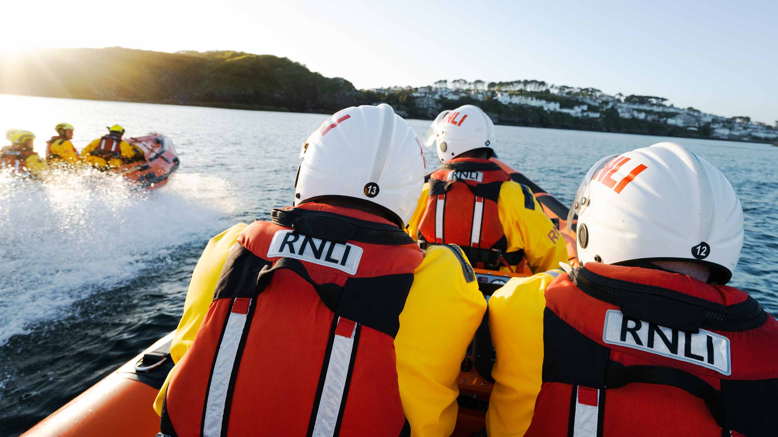Three lifeboat crew members wearing lifejackets and helmets cruise across the water with a D class lifeboat motoring nearby. In the distance, the town of Fowey sits on the horizon. The sun is low in the sky.