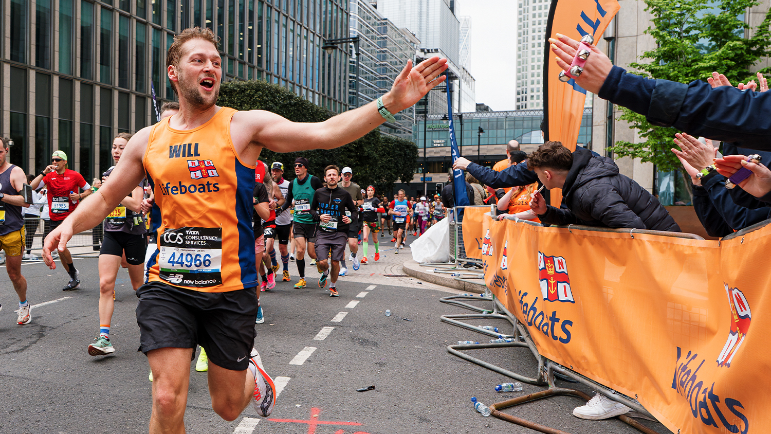 A man running the London Marathon for the RNLI reaches out to high five a woman cheering him on from the sidelines. There are other people running in the background.