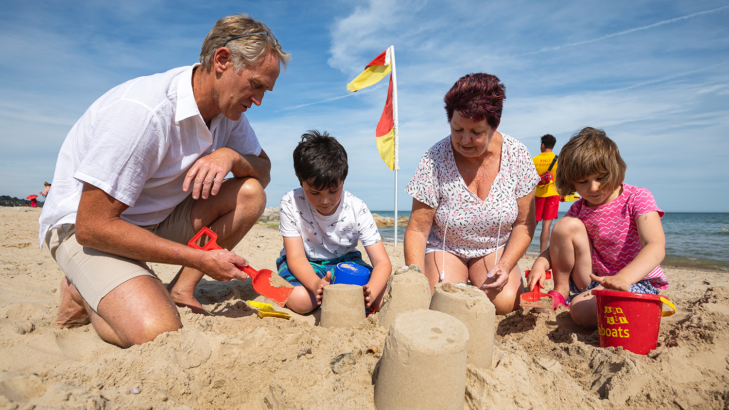 A family of four build sandcastles on the beach, with a lifeguard and a red and yellow flag at the water’s edge
