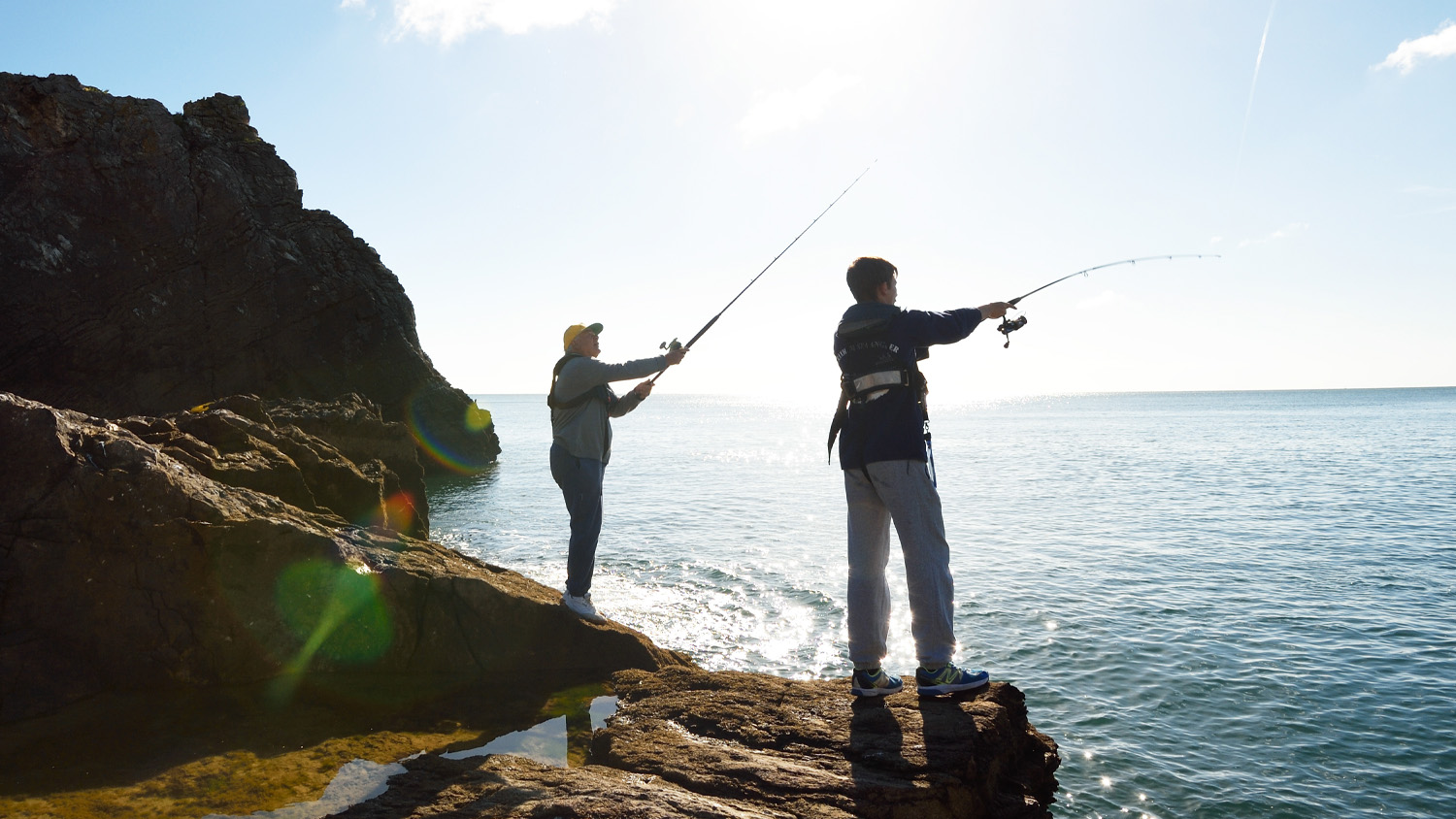 Two people wearing lifejackets fishing from a rock over the sea