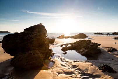Rocks and sand on the beach, with the sea and the sun beginning to set in the background.