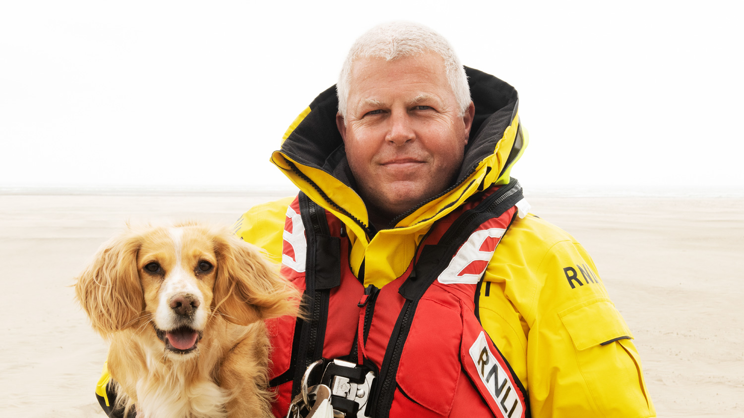 A crew member standing on a sandy beach, holding a dog