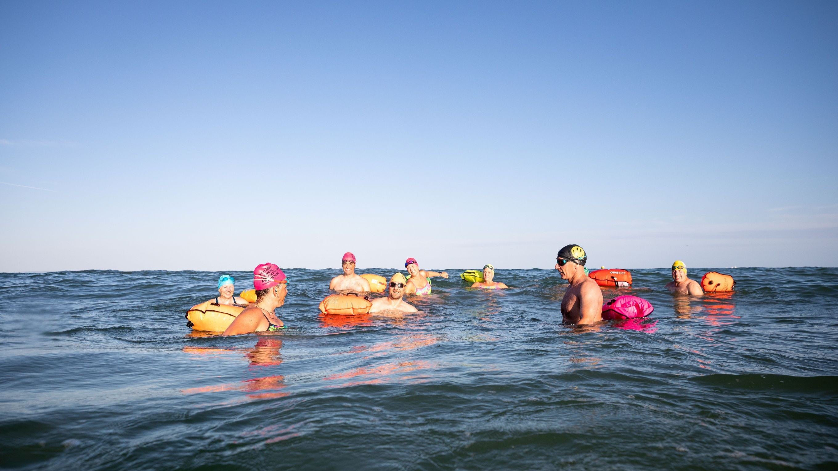 A group of smiling cold water dippers bobbing in a calm sea and wearing colourful swimming hats and tow floats. 