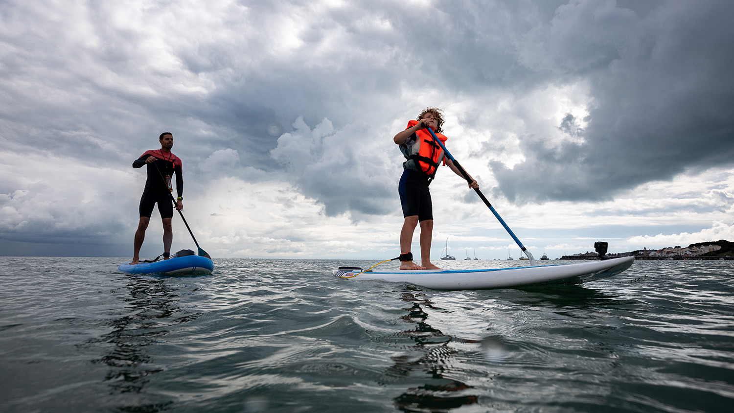 Two paddleboarders, wearing wetsuits and buoyancy aids, paddling on the sea