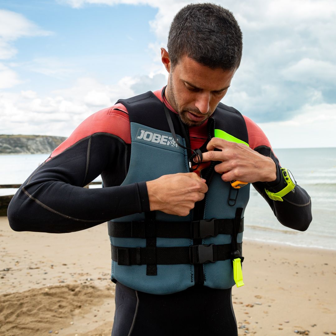 A man is fastening his buoyancy aid while stood on the beach on a sunny, calm day.