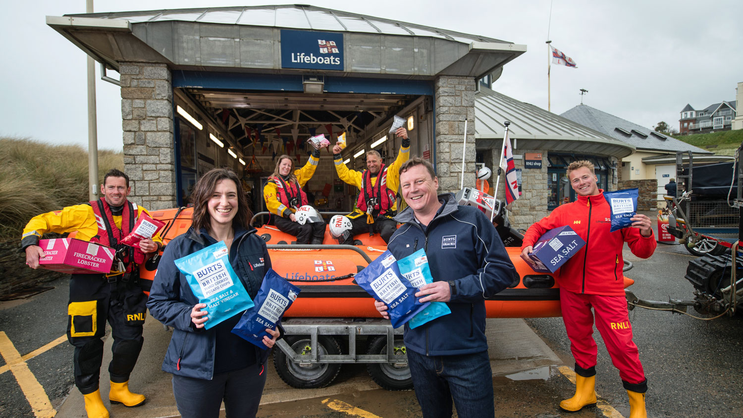 Bude’s D class lifeboat on its trailer outside the boathouse with two lifeboat volunteers sitting inside, one standing next to it on the left, an RNLI lifeguard standing to its right, and Lucy Ashton from the RNLI and Dave McNulty from Burts Snacks standing in front of it. All smiling and holding packs of RNLI-branded Burts Snacks hand-cooked crisps. 