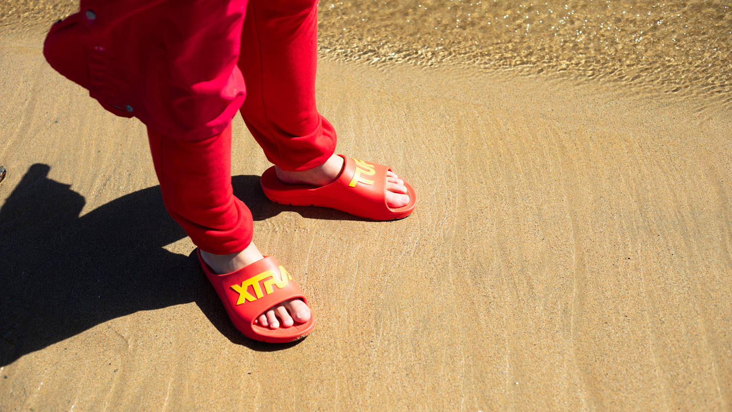 An RNLI lifeguard wearing a pair of bespoke red and yellow sliders from XTRATUF. They’re standing on the sand, with the sea in the background.