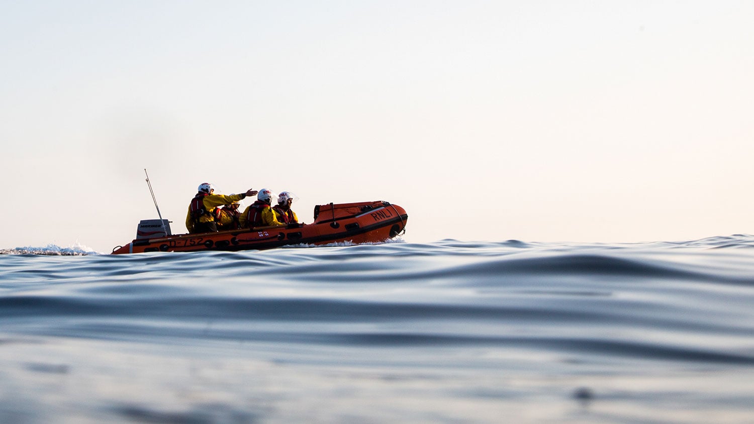 An inshore lifeboat is afloat on the horizon in relatively calm waters underneath a bright white cloudy sky.