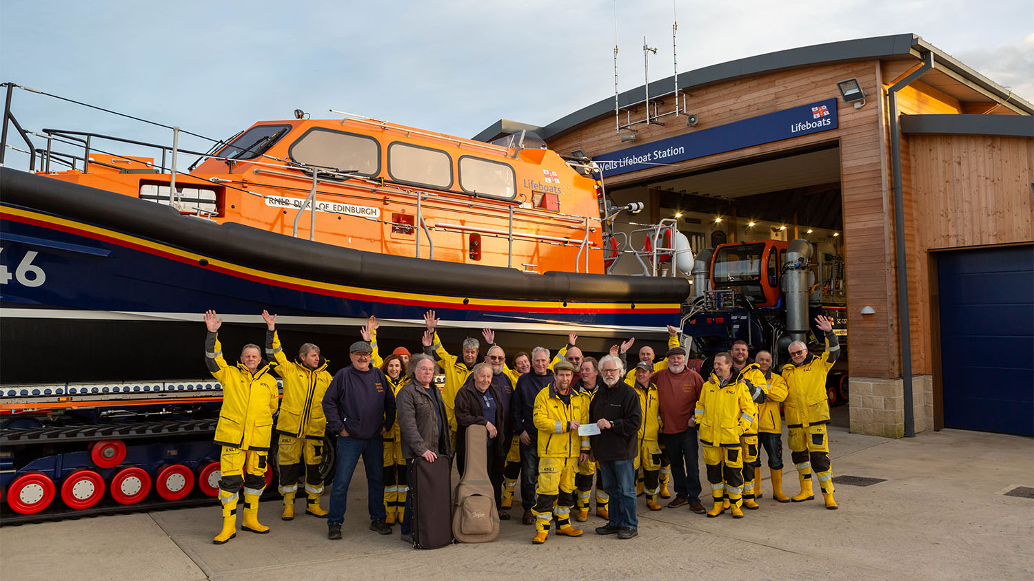 Fundraisers and RNLI crew members stand waving to camera in front of the all-weather lifeboat at Wells Lifeboat Station.