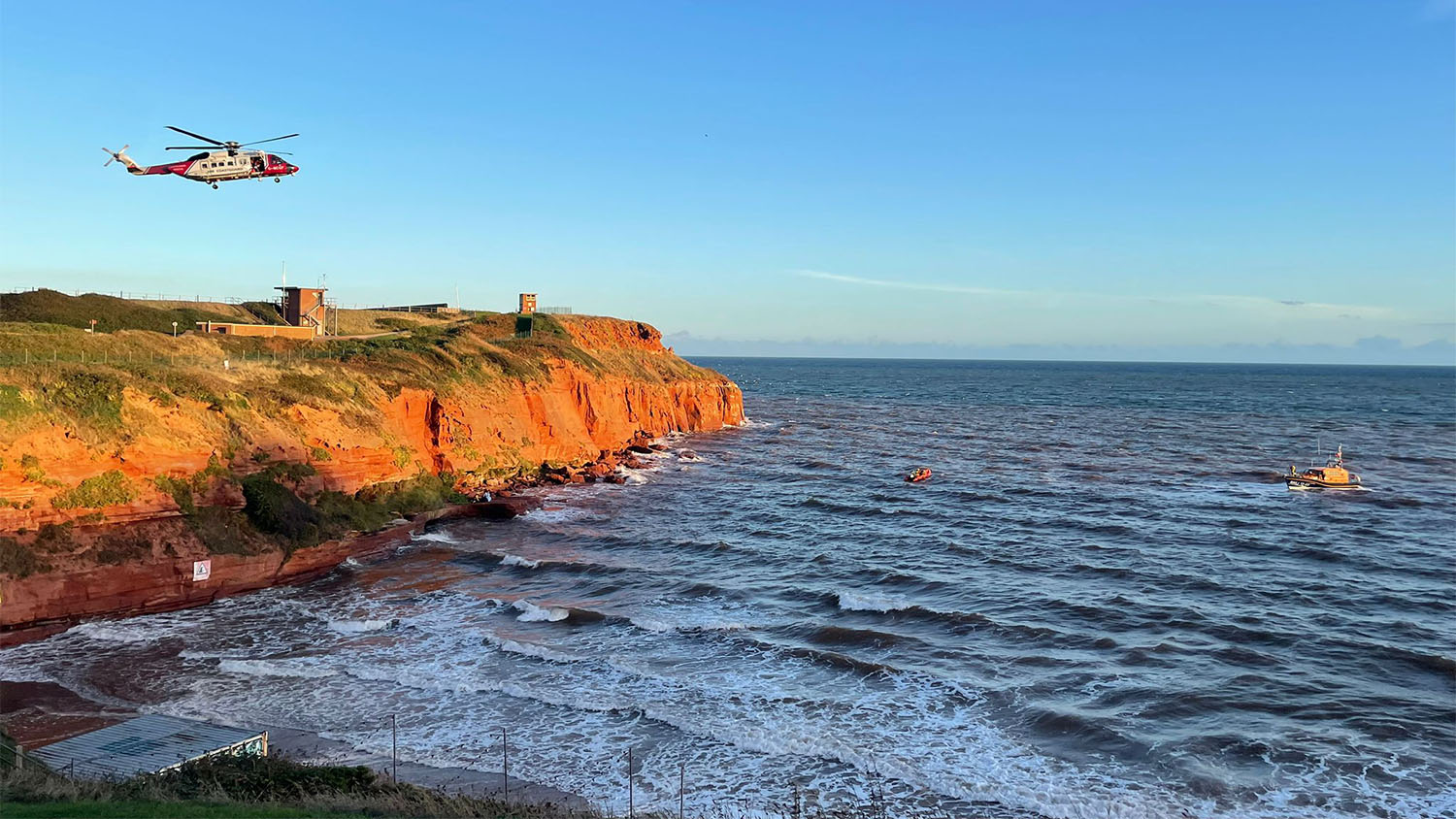 An H.M. Coastguard helicopter hovers over a cliff while two Exmouth RNLI lifeboats get into position in waters near the cliff.