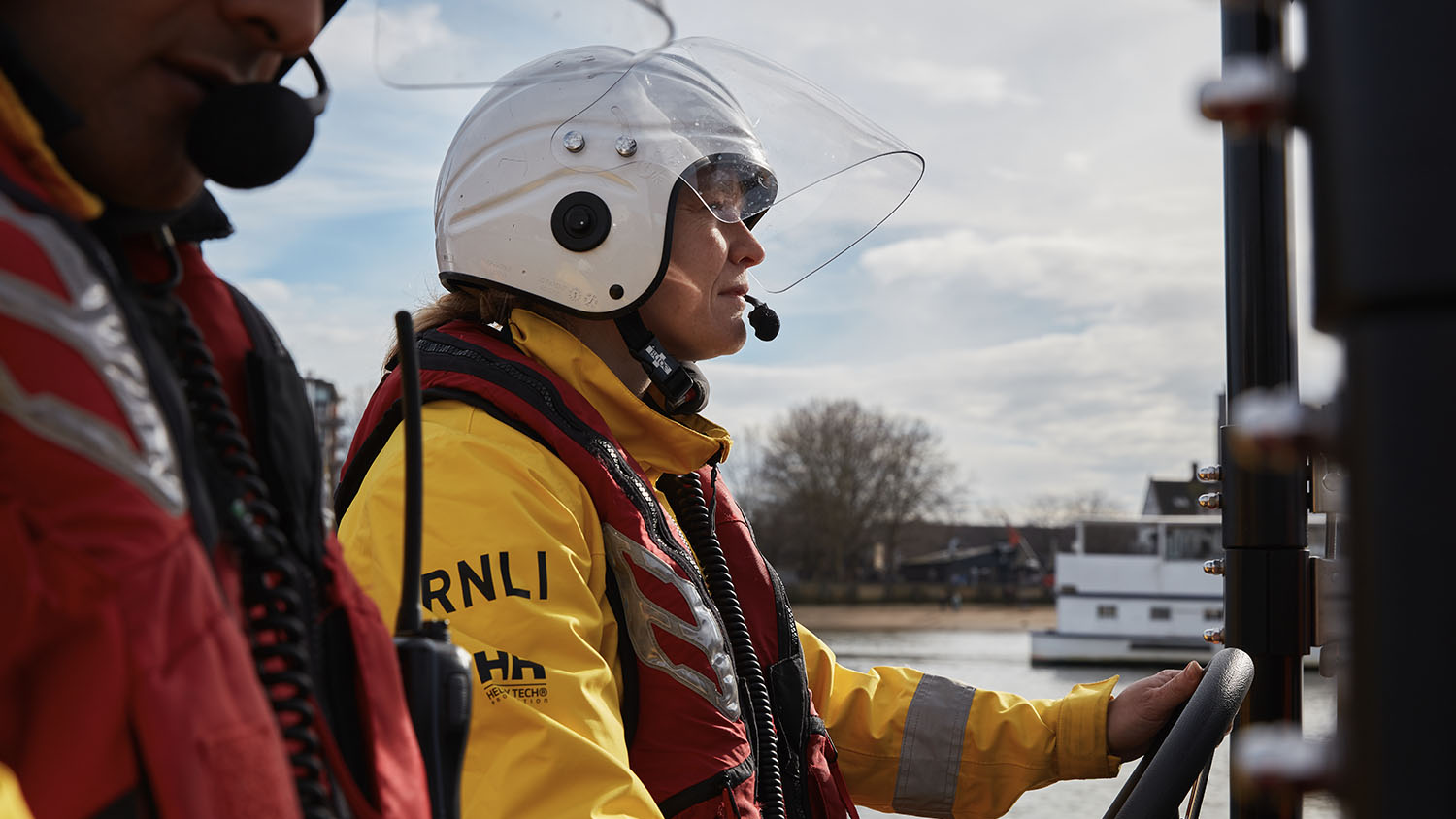 Close-up, side profile of two RNLI E class crew members in crew kit. The female crew member is driving the lifeboat on the River Thames in London.