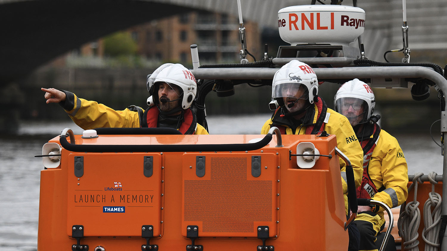 Two crew members at the helm of an E class lifeboat on the River Thames. Two panels show how names will appear on the Thames Launch a Memory lifeboat.