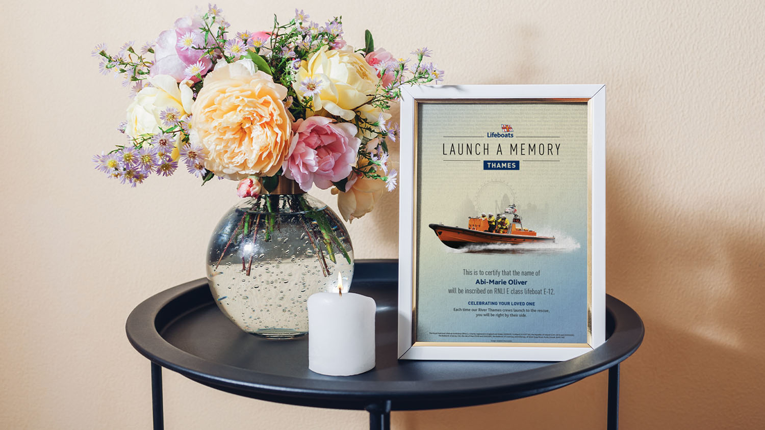 A framed Launch a  Memory certificate on a small round table with a vase of beautiful flowers and a lit candle.