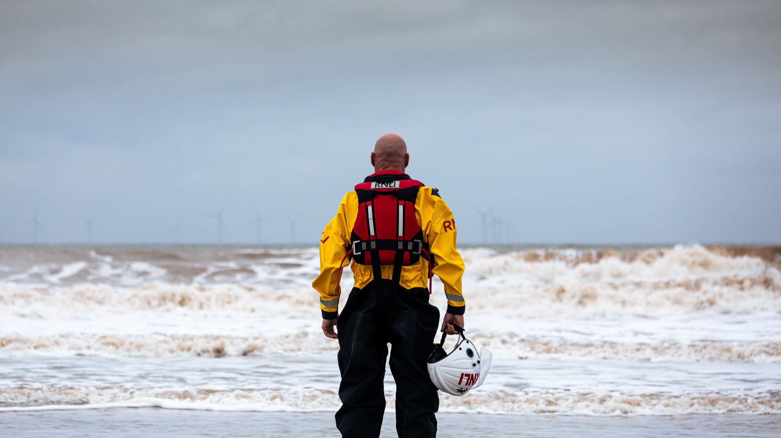 An RNLI crew member stands alone on a beach, staring out to sea