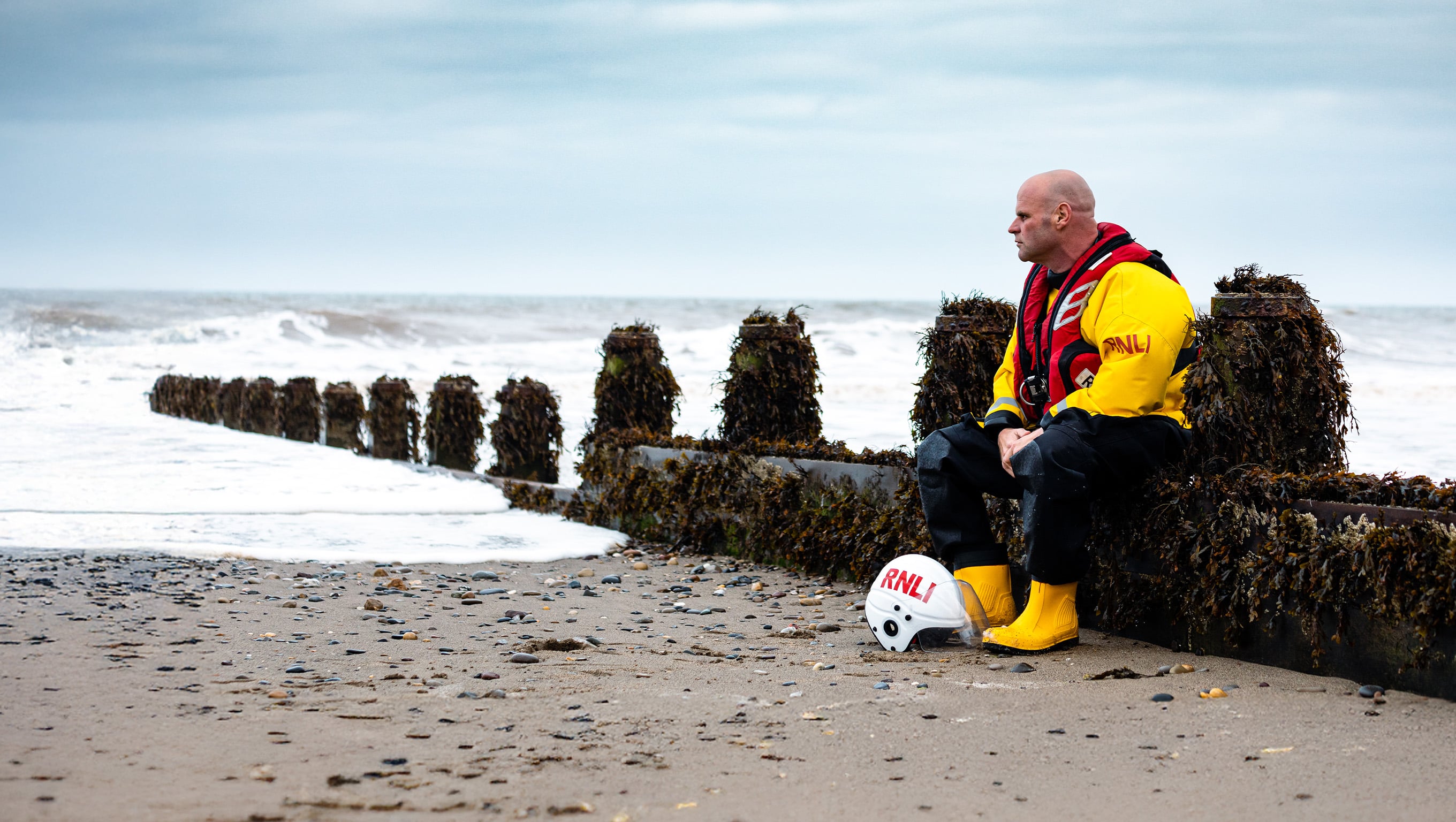 Crew member sitting on a groin on the beach, looking out to sea. 