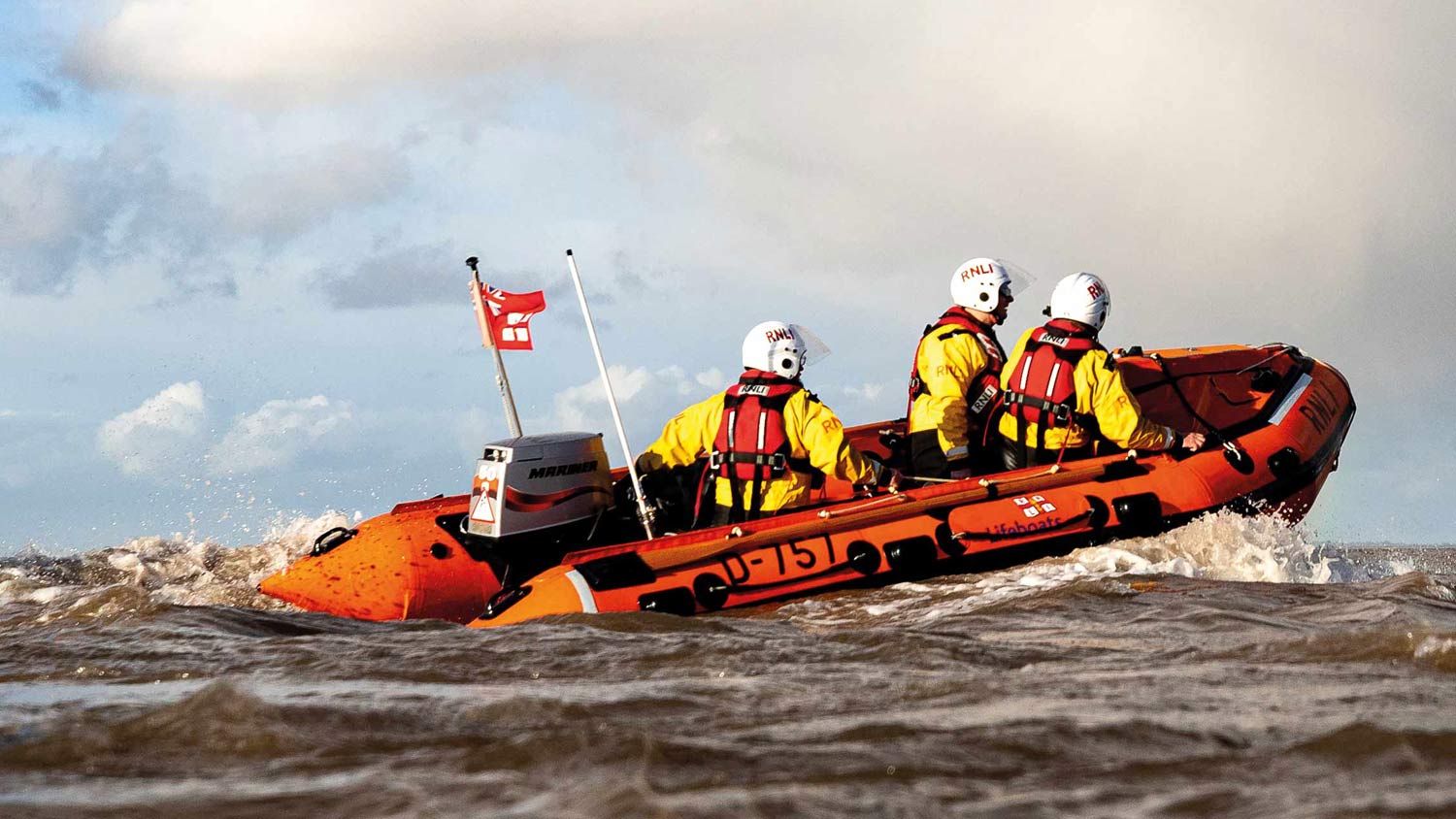Cleethorpes D class lifeboat cruises across the water with three crew members onboard. The crew are wearing yellow inshore lifeboat kit and white helmets. It's a bright day with some cloud on the horizon.