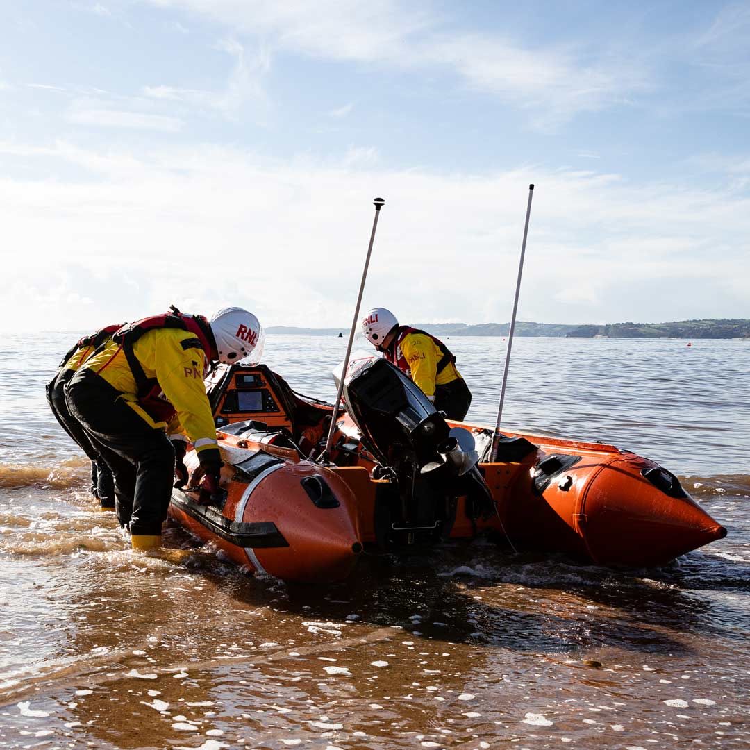 Two lifeboat crew members wearing yellow inshore lifeboat kit and white helmets prepare a D class as it sits at the edge of the surf line. It's a bright sunny day.