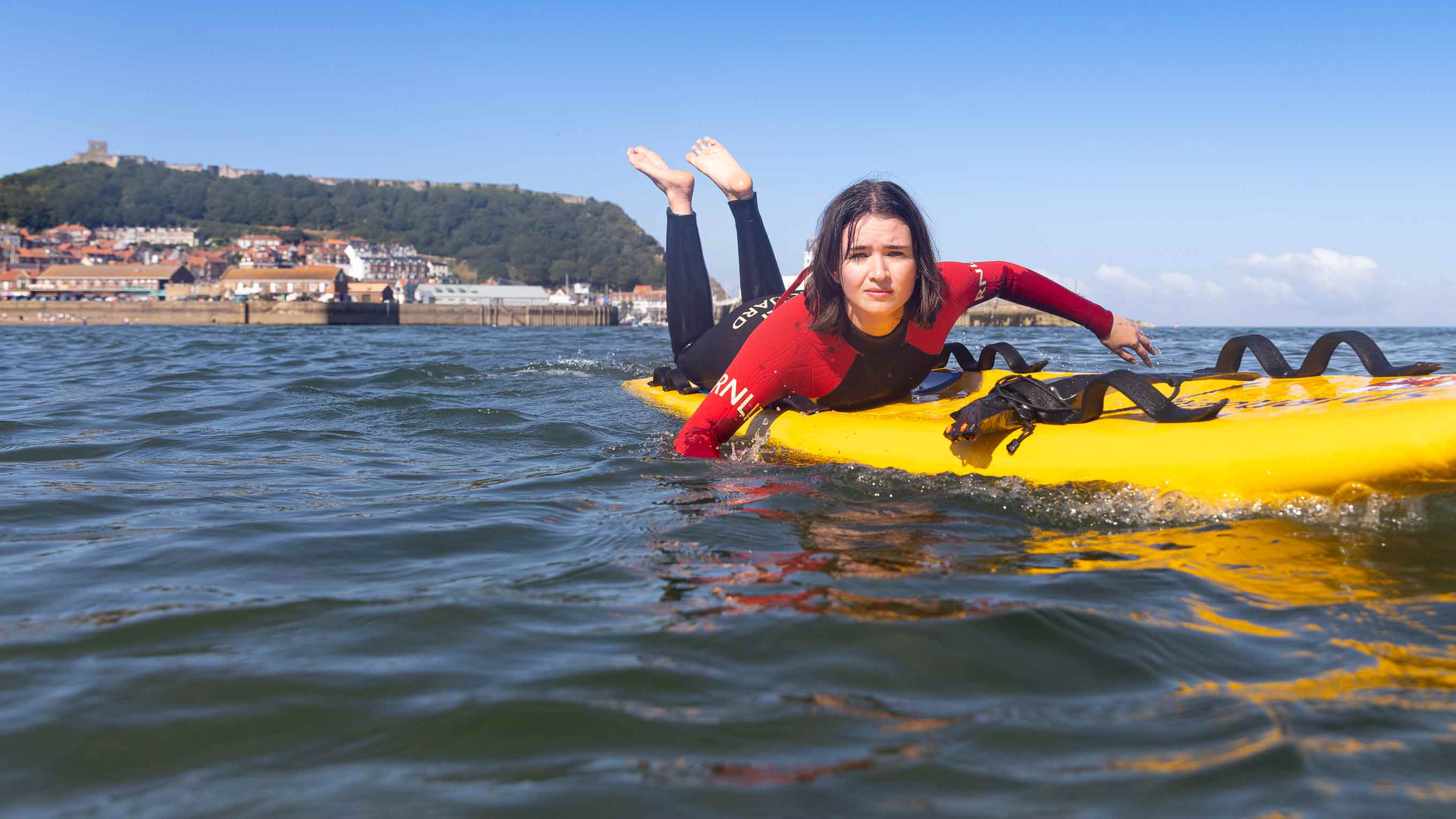A female lifeguard wearing a red and black RNLI wetsuit paddles towards the camera on the back of a bright yellow rescue board