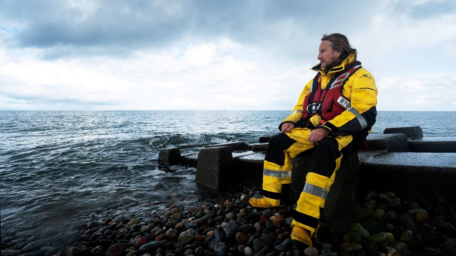 A crew member sits on a beach groyne looking out to sea.